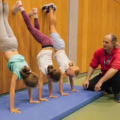 Drei Kinder üben Handstände an der Wand, während ein Trainer der Turngemeinde Landshut aufmerksam zusieht.