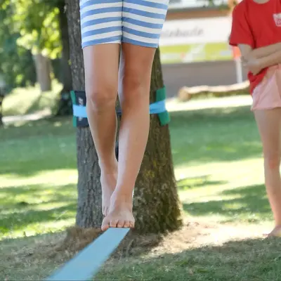 Ein Kind balanciert auf einer Slackline im Park, während ein anderes im TGL-Shirt zuschaut.