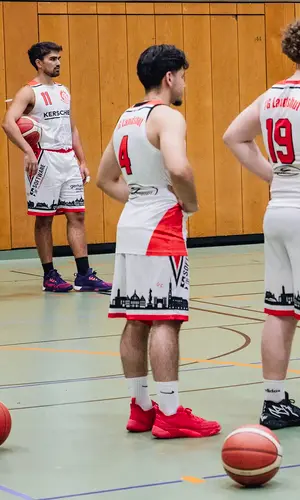 Basketballspieler der TG Landshut bereiten sich in der Halle auf ihr nächstes Spiel vor, während sie gespannt Anweisungen folgen.
