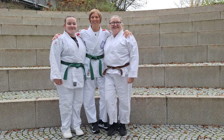 Drei Frauen in Judoanzügen posieren lächelnd auf einer Treppe, stolz ihre Zugehörigkeit zum Turngemeinde Landshut Verein zeigend.