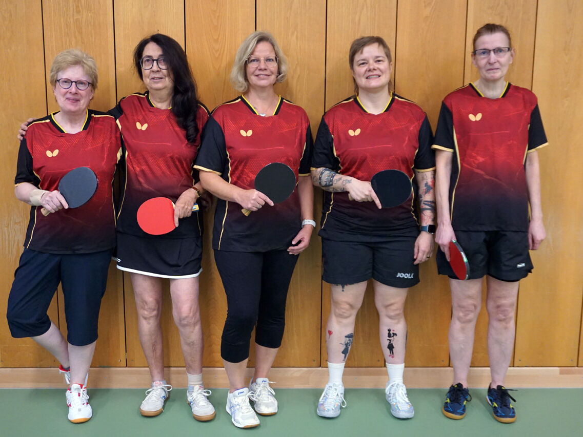 Fünf Frauen in Turngemeinde-Trikots posieren mit Tischtennisschlägern vor einer Holzvertäfelung.