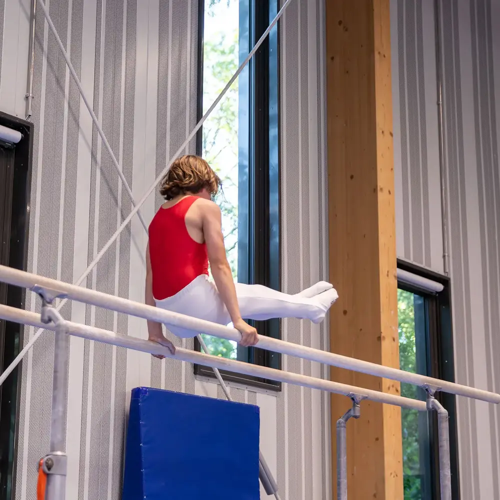 Ein Turner in rotem Trikot zeigt eine Übung am Barren in der Turnhalle des Vereins Turngemeinde Landshut.