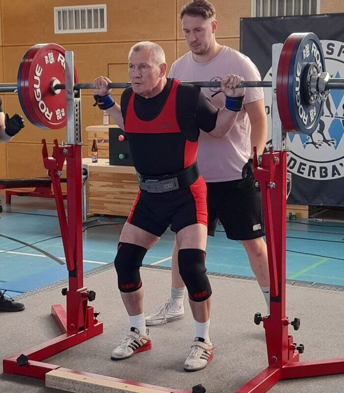 Ein Sportler der Turngemeinde hebt Gewichte, unterstützt von einem Trainer in einer Sporthalle.