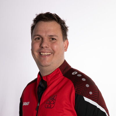 Ein Mann lächelt in einer roten TGL-Jacke, repräsentativ für den Sportverein Turngemeinde Landshut.