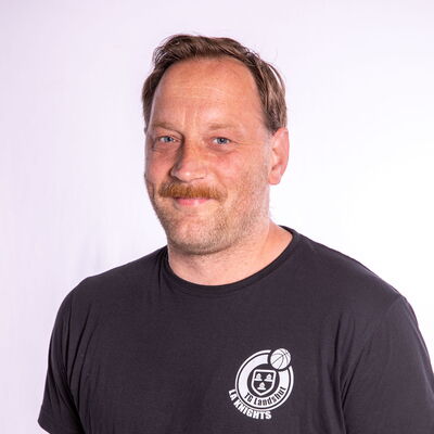 Ein Mann mit Bart trägt ein schwarzes T-Shirt mit einem Basketball-Logo, repräsentativ für den Verein Turngemeinde Landshut.