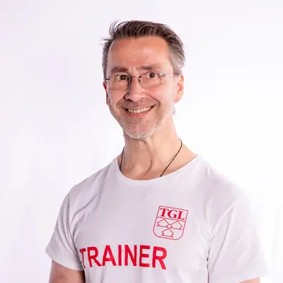 Ein lächelnder Trainer in einem TGL-Shirt steht vor einem weißen Hintergrund und repräsentiert den Sportverein.