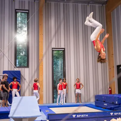Ein Turner des Vereins Turngemeinde Landshut führt einen Salto auf dem Trampolin aus, während andere zuschauen.