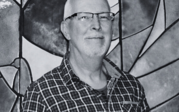 Ein Mann mit Brille lächelt vor einem kunstvoll gestalteten Glasfenster, das an die Turngemeinde Landshut erinnert.