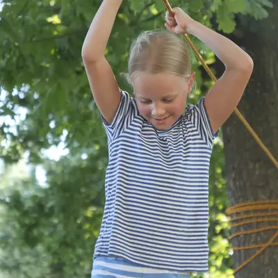 Ein Mädchen in einem gestreiften Shirt schwingt fröhlich an einem Seil im Freien, was an einen Sport im Turngemeinde-Stil erinnert.