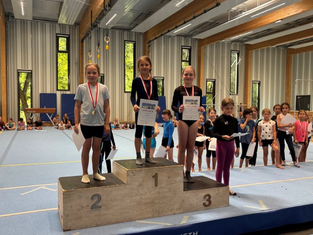 Drei junge Turnerinnen stehen auf einem Siegerpodest und präsentieren stolz ihre Urkunden beim Turnvereinwettbewerb.