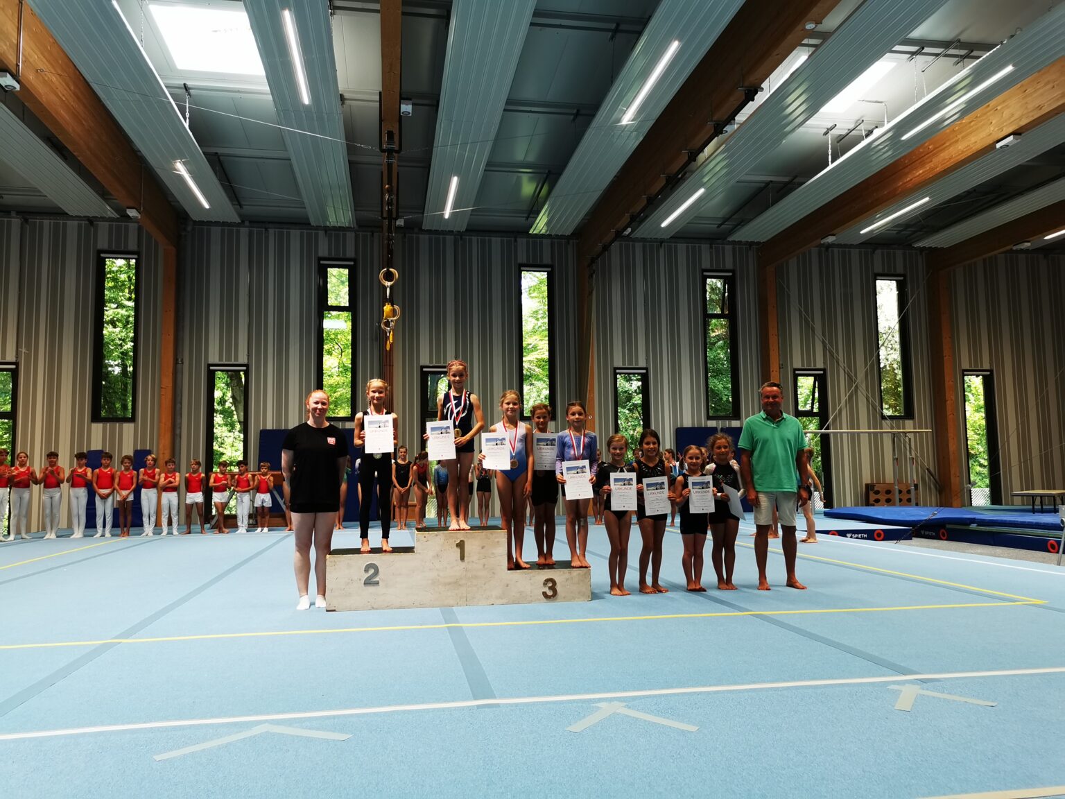Mädchen stehen auf einem Podest in einer Turnhalle und halten Urkunden, während sie bei einem Turngemeinde-Wettbewerb geehrt werden.